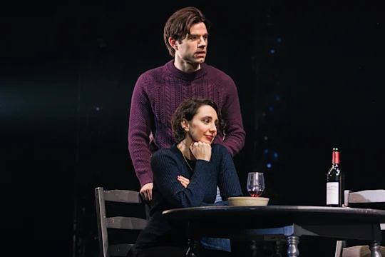 A man stands behind a seated woman at a table set with a wine bottle, wine glass, and bowl. Both appear thoughtful and are dressed in dark sweaters.