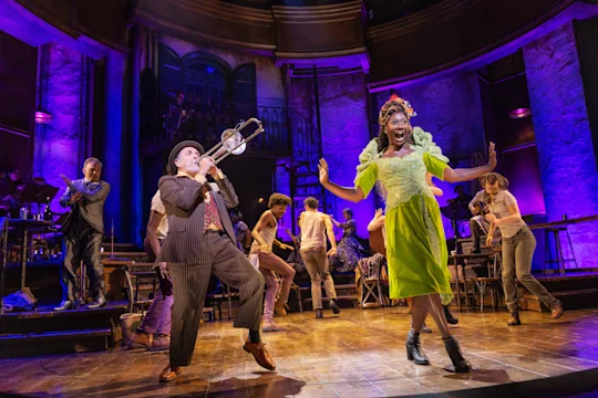 Actors perform on a brightly lit stage; one plays a trombone while another, wearing a green dress, dances. The set has purple lighting and several background performers.