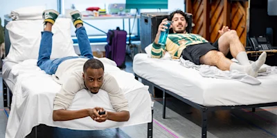 Two men are relaxing on separate twin beds in a room; one is lying on his stomach using a TV remote, and the other is lying on his back holding a water bottle.