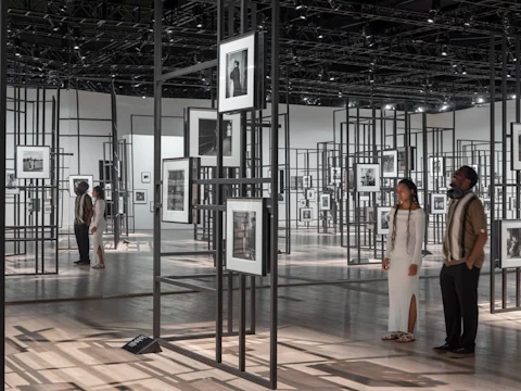 People view black-and-white photographs suspended in frames throughout a modern art gallery with a grid-like metal structure.