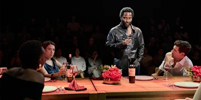 A man stands while holding a wine glass at a table with three seated people, bottles of wine, and flowers, as an audience watches in the background.