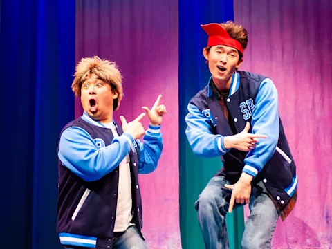 Two performers on stage wear blue jackets and red bandanas, striking playful poses against a colorful background.