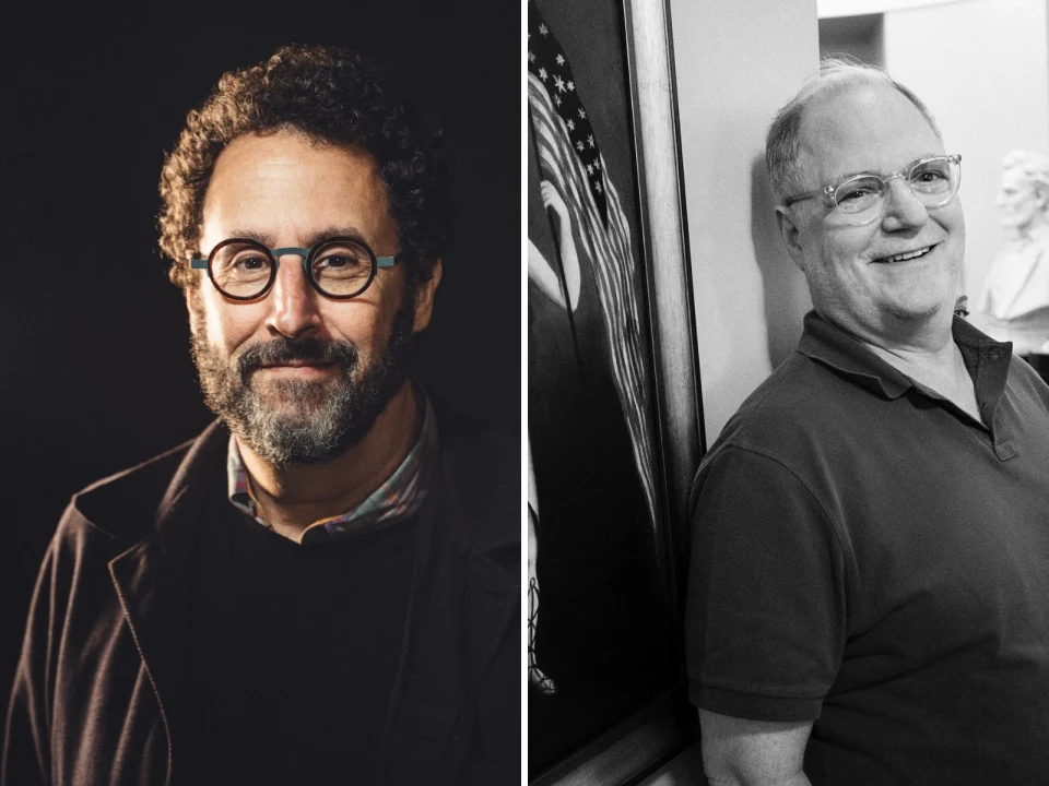 Two men are pictured: the left one with curly hair, glasses, and a beard, wearing a dark jacket; the right one with short hair and glasses, smiling, in a casual shirt, standing by a wall.