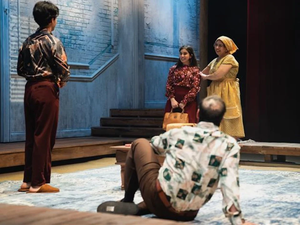 Four people are on stage in a theatrical performance; two women stand together, one man sits on the floor facing them, and another man stands with his back to the camera.