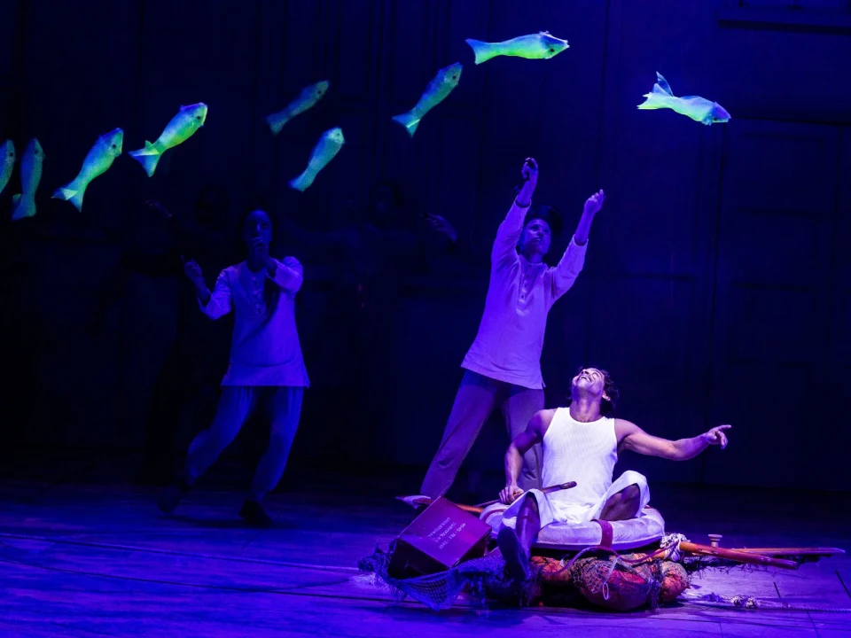 A stage performance with a person sitting on a bed of cushions pointing towards floating fish, surrounded by other performers in a dimly lit, blue-purple setting.