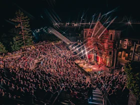 A large crowd sits in an outdoor amphitheater at night, watching a live performance on a brightly lit stage next to a historic building.