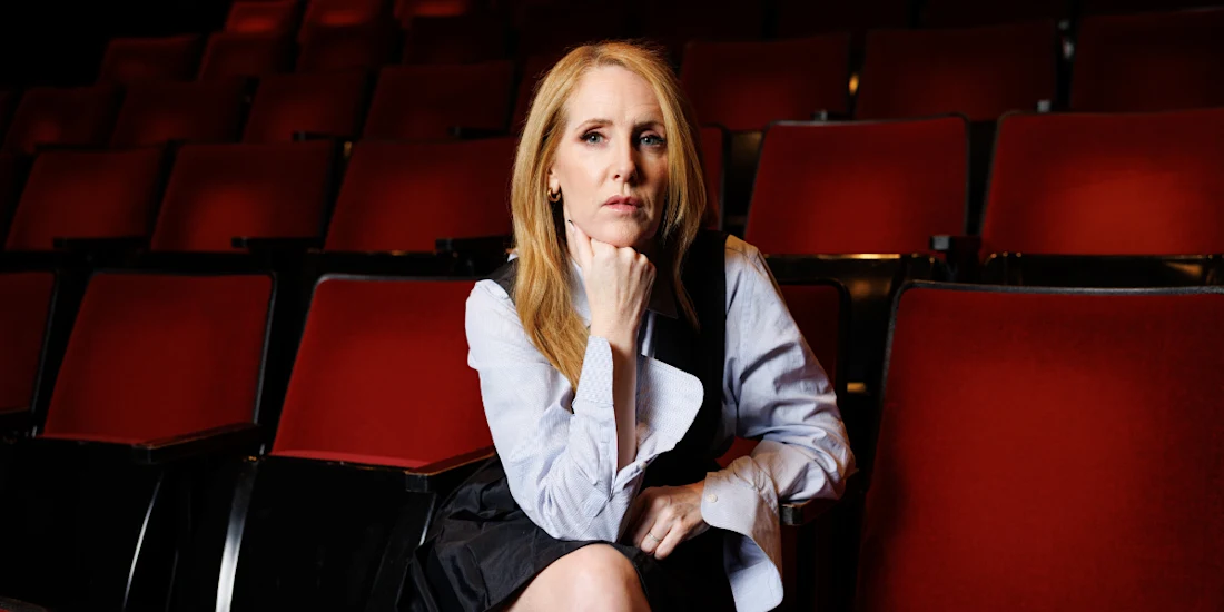 A woman with long blonde hair sits in a mostly empty theater, resting her chin on her hand, wearing a light blue blouse and dark dress. Red seats fill the background.