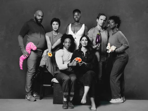 Cast sitting and standing in a black and white photo holding selectively colored objects like a grapefruit, eggplant, white rabbit, and more.