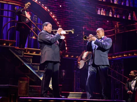 Musicians perform on stage, with two men playing trumpets at the forefront. Others play instruments in the background amidst a vibrant theater setting.