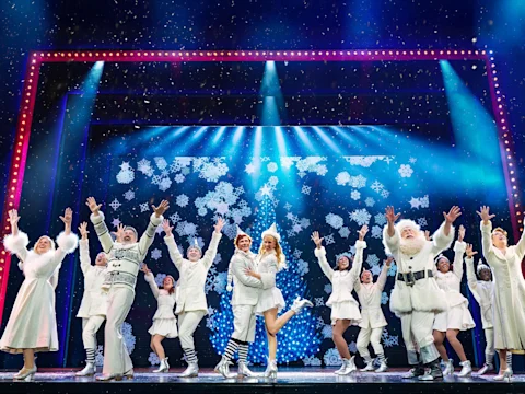 Performers dressed in white costumes dance and pose on a brightly lit stage with snowflake decorations and sparkling lights.