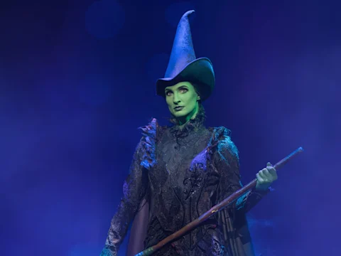 A person in a green face makeup and ornate black costume with a tall pointed hat stands holding a staff, surrounded by blue stage lighting and theatrical smoke.