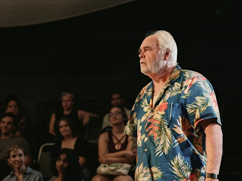 An older man in a floral shirt stands and faces forward, with several seated people watching him in the background.