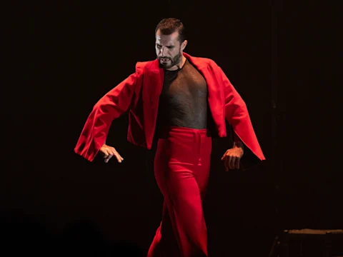 A dancer in a red jacket and red pants performs on stage with dramatic arm movements against a dark background.