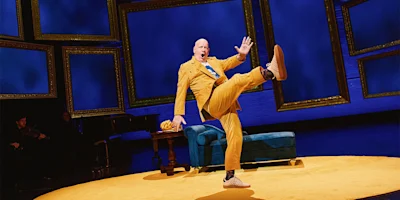A bald man in a yellow suit performs an exaggerated kick on stage, with picture frames, a blue couch, and a table in the background.