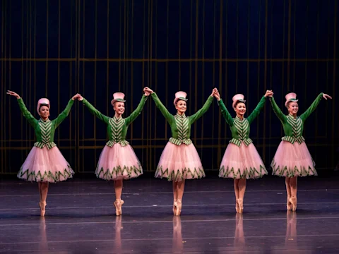 Five ballet dancers in green jackets and pink tutus stand on stage in a line, holding hands with arms raised, wearing matching hats and performing en pointe.
