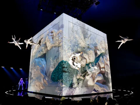 Performers in white costumes are suspended in air around a large cube with nature-themed projections, inside a dimly lit venue.