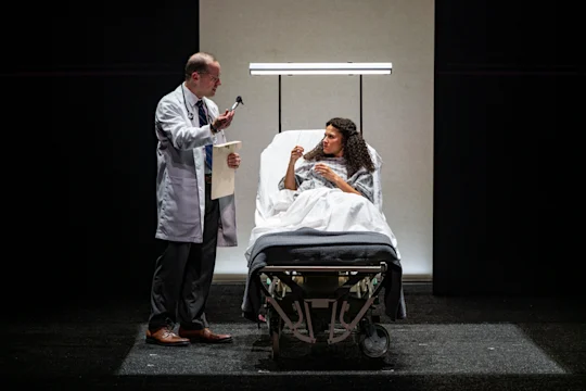 A doctor stands beside a hospital bed, speaking to a woman in a hospital gown who is sitting up and gesturing with her hand, in a clinical setting under bright lighting.