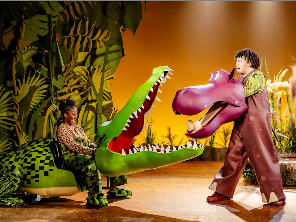 Three people manipulate bird puppets near a large crocodile puppet with its mouth open, surrounded by tall grasses and plants on a stage.