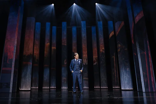 A person in a suit stands alone on a stage with tall vertical panels and overhead spotlights illuminating the scene.