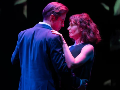 Two people in formal attire dance closely while looking into each other's eyes on a dimly lit stage.
