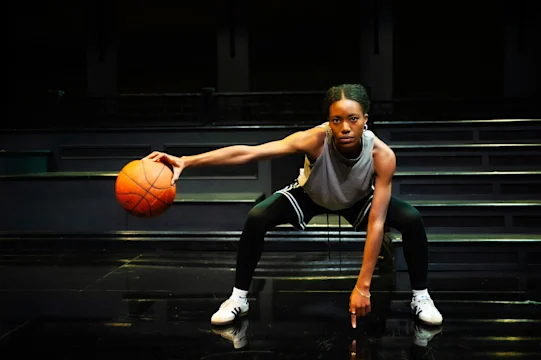 A basketball player in athletic wear dribbles a basketball low to the ground on an indoor court, looking forward intently.