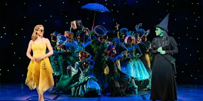 Two women stand on stage in costume, one in a yellow dress and one in black with green skin, while a group in elaborate green outfits gathers behind them under stage lights.