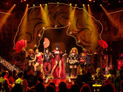 Performers in vibrant costumes dance on a stage with a colorful, wavy backdrop and dramatic lighting, while an audience watches.