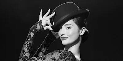 Woman in lace dress poses with one hand holding the brim of a black top hat, looking at the camera and smiling against a dark background.