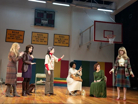 A group of six women stands on a gymnasium stage, with one woman in the foreground speaking. A scoreboard and basketball hoop are visible in the background.