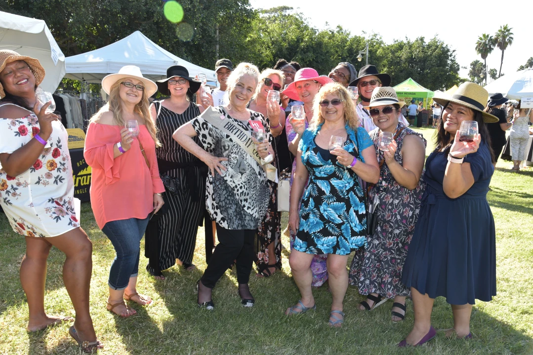 Group of women in dresses and hats, smiling and holding wine glasses at an outdoor event, with tents in the background.