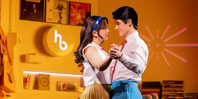 A man and woman in retro-style clothing dance closely in a warmly lit room with yellow walls, records, and vintage decor in the background.