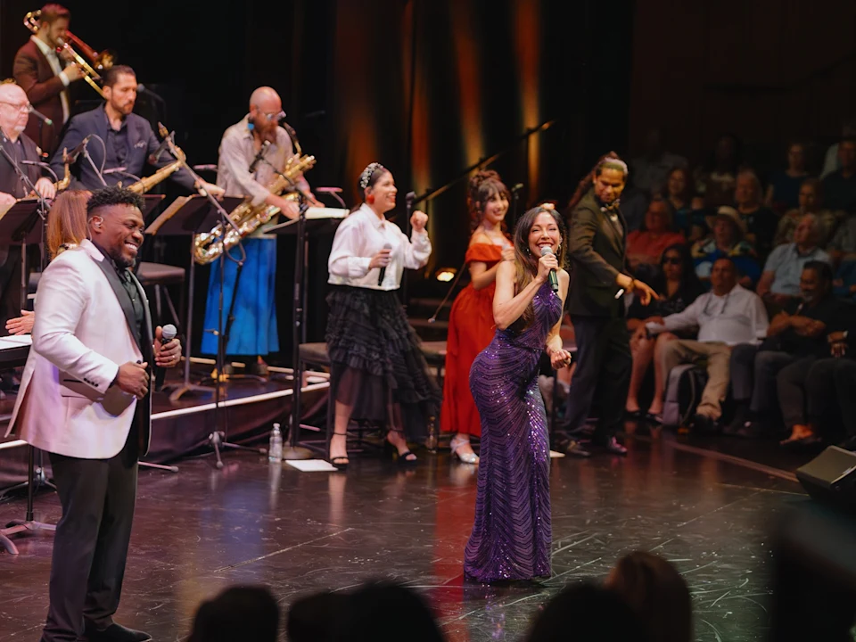 A woman sings into a microphone on stage with a band and several other performers, while an audience watches from the seats.