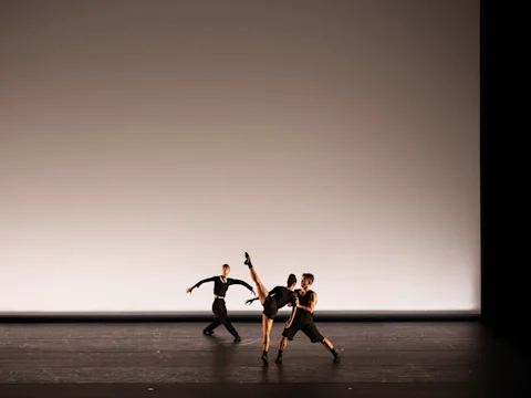 Three dancers perform on stage; one lifts a leg high, supported by another, while the third moves in the background. The backdrop is plain, and they are dressed in black.