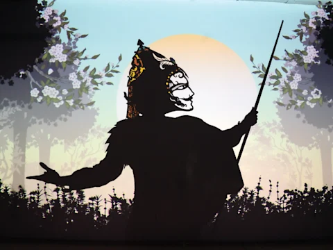 Silhouette of a figure holding a stick, wearing a decorative mask, set against a backdrop of trees and a circular light source.