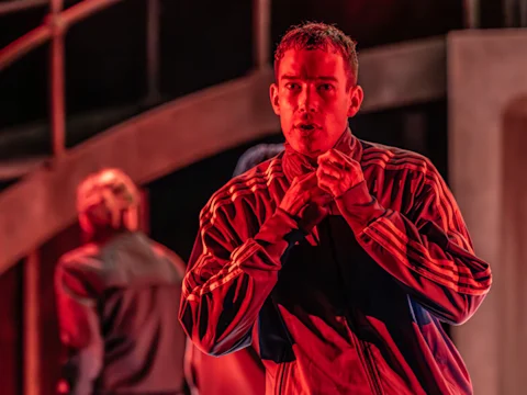 Photo of David Shields as Jacob in a red tracksuit jacket and intense expression in Punch on Broadway, New York City.