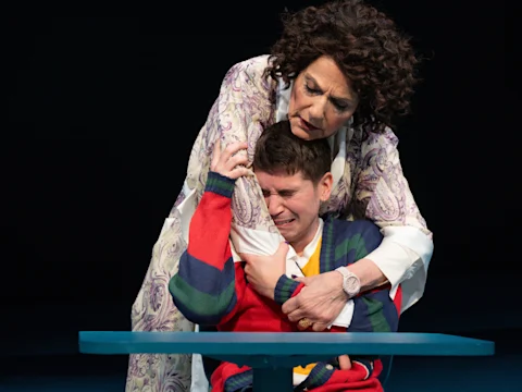 A woman hugs a crying young person who is sitting at a blue table. Both appear emotional, and the scene is set against a dark background.