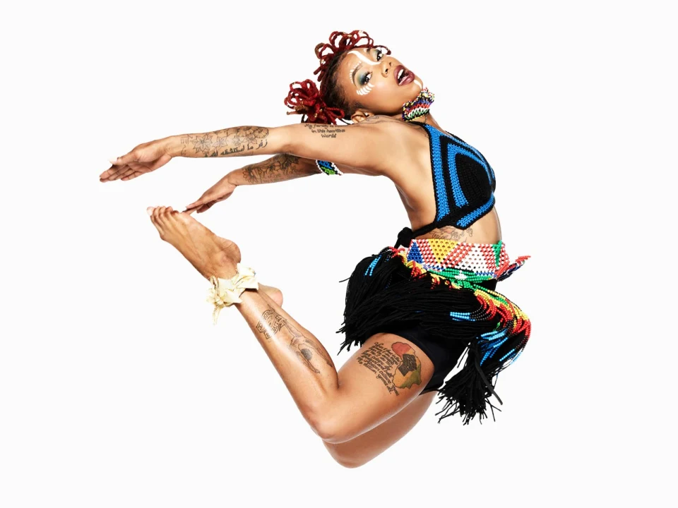 Person with red braided hair and tattoos, wearing a colorful beaded outfit, captured mid-jump with arms and legs extended gracefully against a white background.