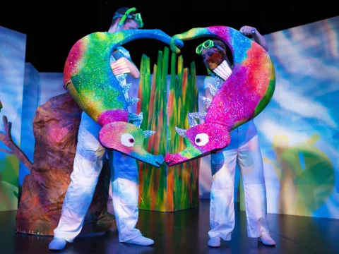 Two performers dressed in white hold large, colorful chameleon puppets in the shape of a heart on stage.