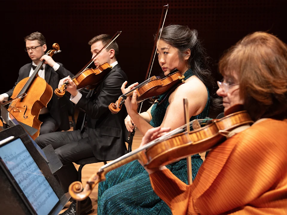 Four musicians perform in a string quartet, playing cello, viola, and violins on a stage; sheet music is visible in the foreground.
