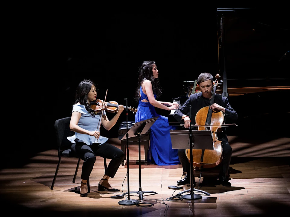 Three musicians perform on stage: a violinist seated left, a pianist in a blue dress center, and a cellist seated right, each playing their respective instruments under stage lighting.