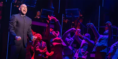 A man stands at the front of a stage, singing passionately, while performers in suits pose dramatically behind him under colorful stage lighting.