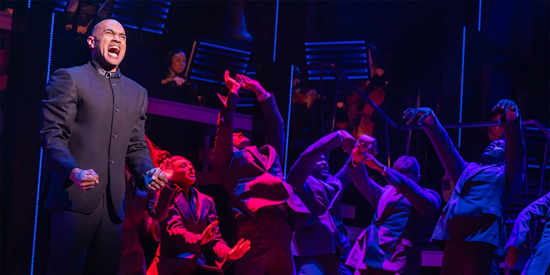 A man stands at the front of a stage, singing passionately, while performers in suits pose dramatically behind him under colorful stage lighting.