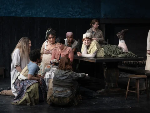 A group of theater actors in costume sits and stands around a wooden table on stage, engaged in conversation. One actor is laying on the table, while others gather closely, looking at something.