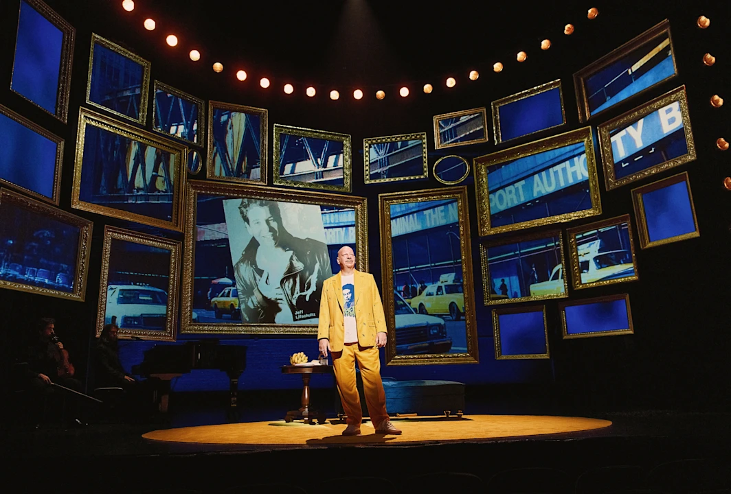 A performer in a yellow suit stands center stage, surrounded by multiple framed screens displaying urban images and taxis under theatrical lighting.