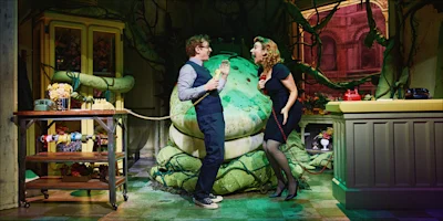 Two actors stand in front of a large green plant prop on stage, holding telephones and smiling, surrounded by floral and kitchen set pieces.