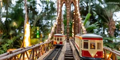 Two model trains cross a miniature wooden bridge decorated with string lights, surrounded by lush greenery.