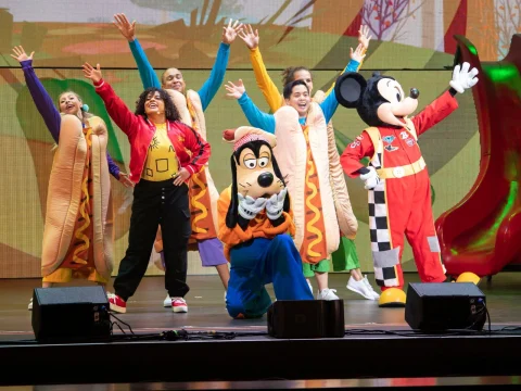 Performers and characters dressed in colorful costumes, including hot dogs, Goofy, and Mickey Mouse, pose energetically on stage during a lively show.