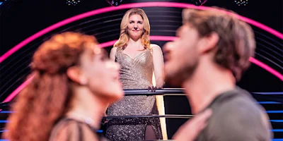 A woman in a shimmering dress stands on a balcony, watching a man and woman in the foreground who appear to share an intimate moment.