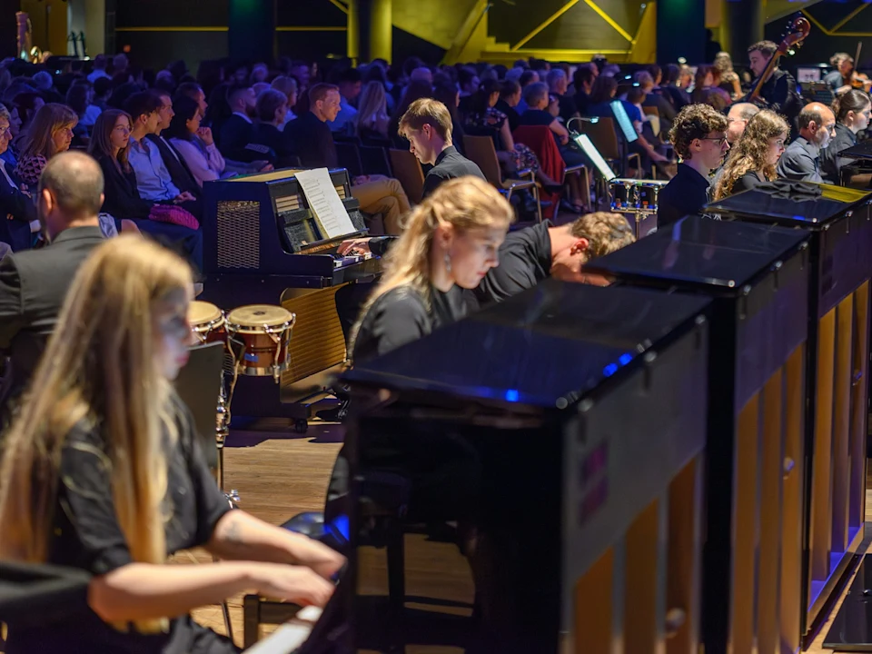 Musicians play pianos and other instruments in an orchestra, with an audience seated in the background in a concert hall.