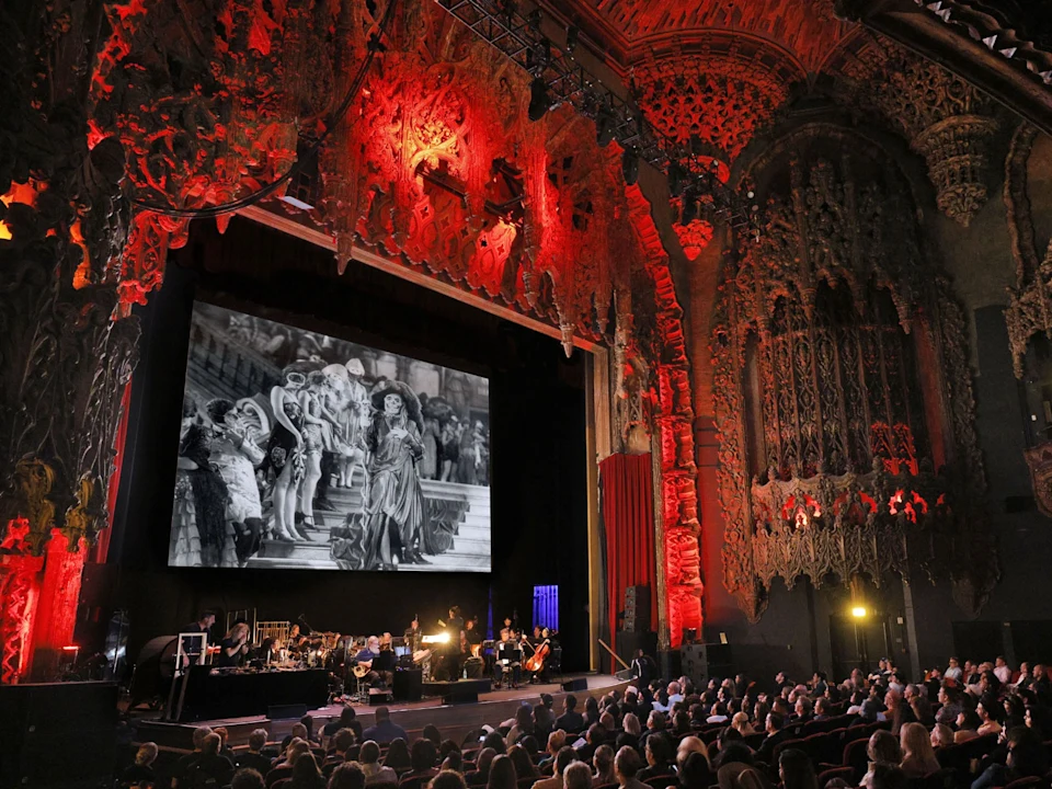 A large audience watches a black-and-white film projected on a theater screen, with musicians performing below and ornate red-lit architecture surrounding the stage.